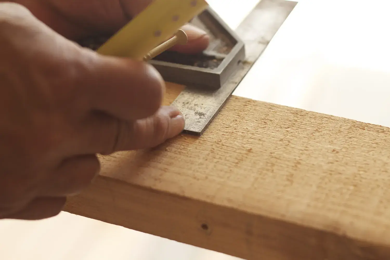 A hand holding a combination square against the edge of a board to mark a line