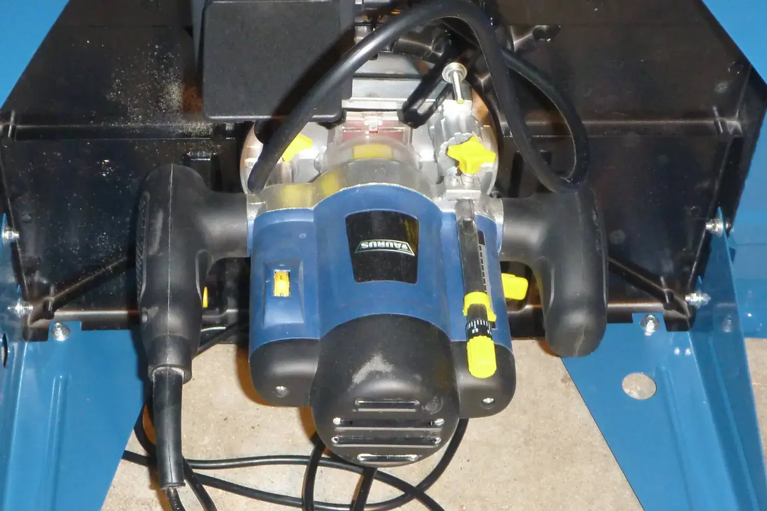 A plunge router mounted upside down under a router table, viewed from below